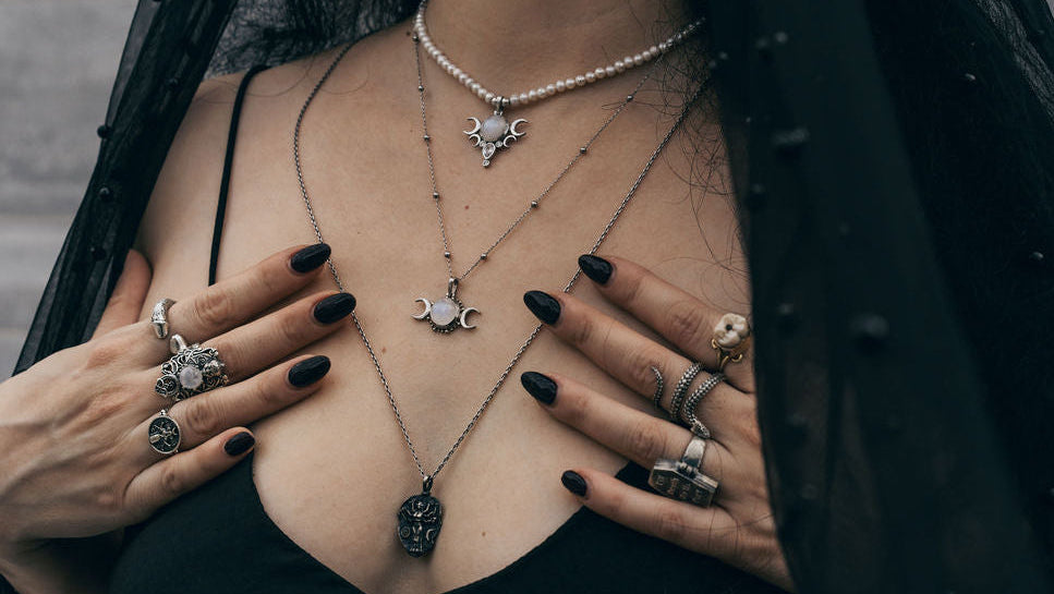 Person wearing multiple necklaces and rings with dark nail polish, against a dark background.