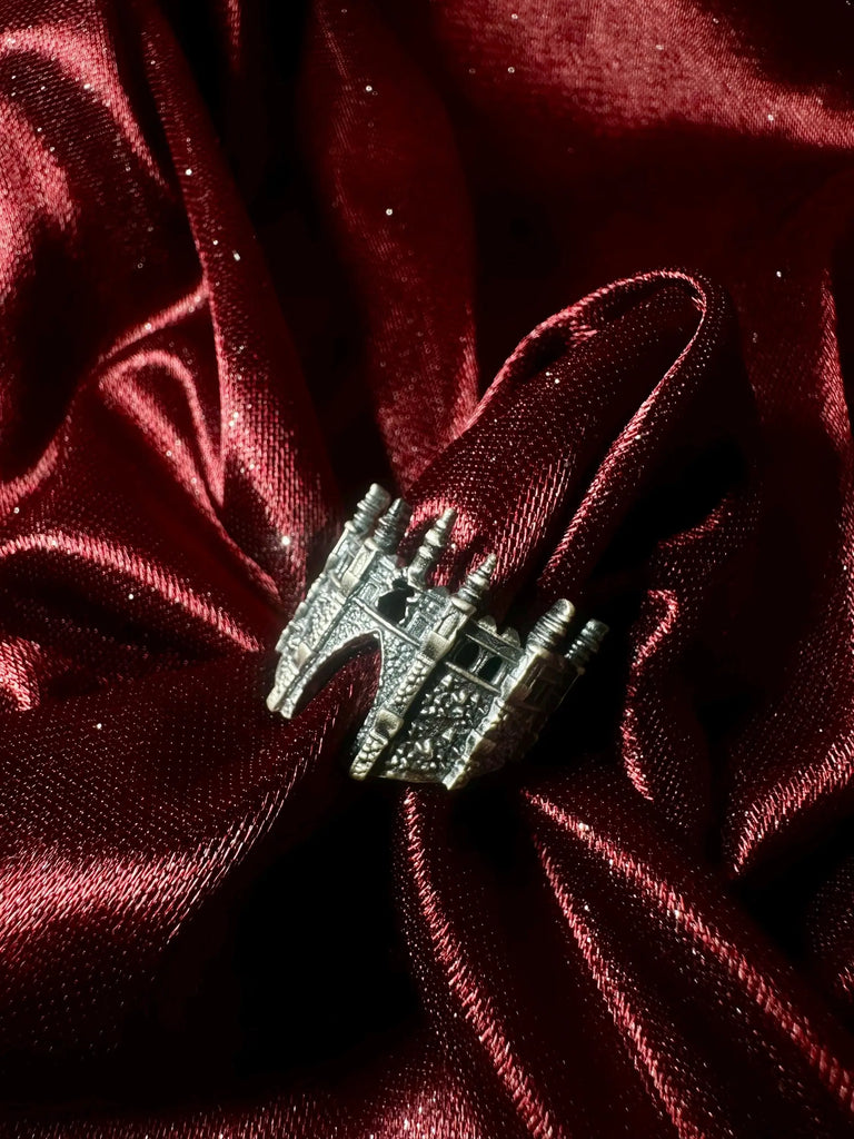 May include: A silver-colored ring with a castle-like design. The ring features multiple turrets and architectural details. It is set against a deep burgundy, textured fabric, creating a contrast between the metallic ring and the rich background. The ring appears to be made of metal.